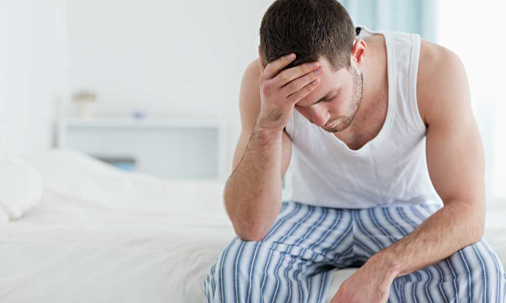Los síntomas de un tumor hipofisario: hombre en pijama sujetándose la cabeza con una mano en el borde de la cama.