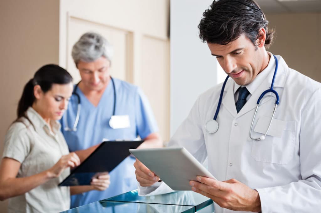 Médico de una clínica masculina de baja T sosteniendo una tableta digital. Un colega y la recepcionista hablan en el fondo.