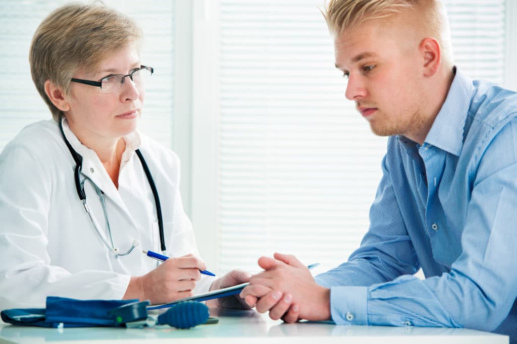 A female doctor wearing a white lab coat and glasses discusses hypothyroidism symptoms with a male patient who has blonde hair and is wearing a blue shirt.