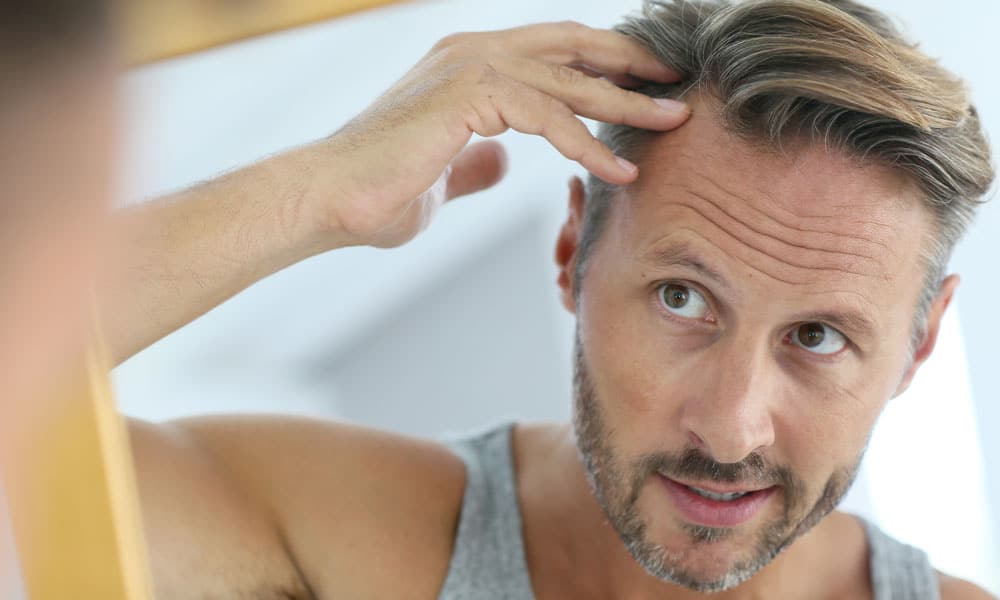 Un hombre comprobando la caída del cabello en el espejo. Muchos hombres temen perder el cabello como consecuencia de la TRT, pero no es así exactamente como se relacionan la testosterona y la caída del cabello. Más información.