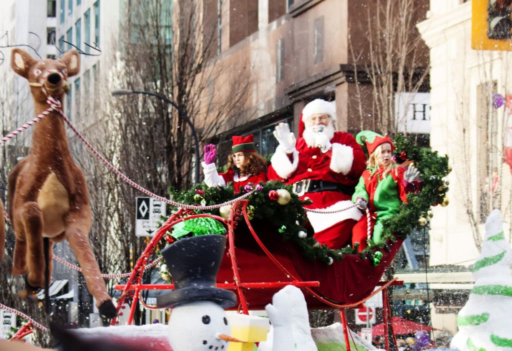Una carroza que transporta a Papá Noel y algunos elfos avanza por una calle de la ciudad. Un muñeco de nieve saluda en primer plano. El desfile de Navidad es uno de los eventos navideños que se celebran cada año en Lewisville.