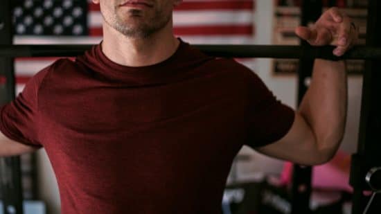 Un hombre levantando pesas en el gimnasio de su casa con una bandera americana de fondo. La terapia de reemplazo de testosterona no es un atajo para la musculación, pero puede ayudar si usted sufre de testosterona clínicamente baja. Más información.