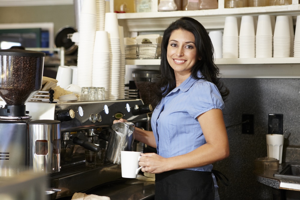 Una mujer con camisa azul que trabaja en una cafetería de Flower Mound saca una taza de café de una gran máquina de café.