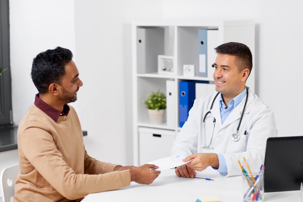 Un paciente con un jersey de color canela y un médico están sentados en una mesa blanca en una consulta médica blanca, posiblemente una clínica de baja testosterona en Dallas-Fort Worth.