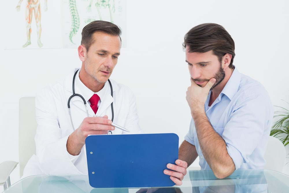 A doctor and a patient sit at a glass table while looking at exam data on a blue clipboard. The doctor gestures with his pen, possibly explaining hypothyroidism treatment available in Dallas- Fort Worth.