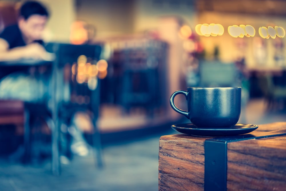 Black coffee mug on a saucer sits on the corner of a rustic wood table in a Prosper cafe.