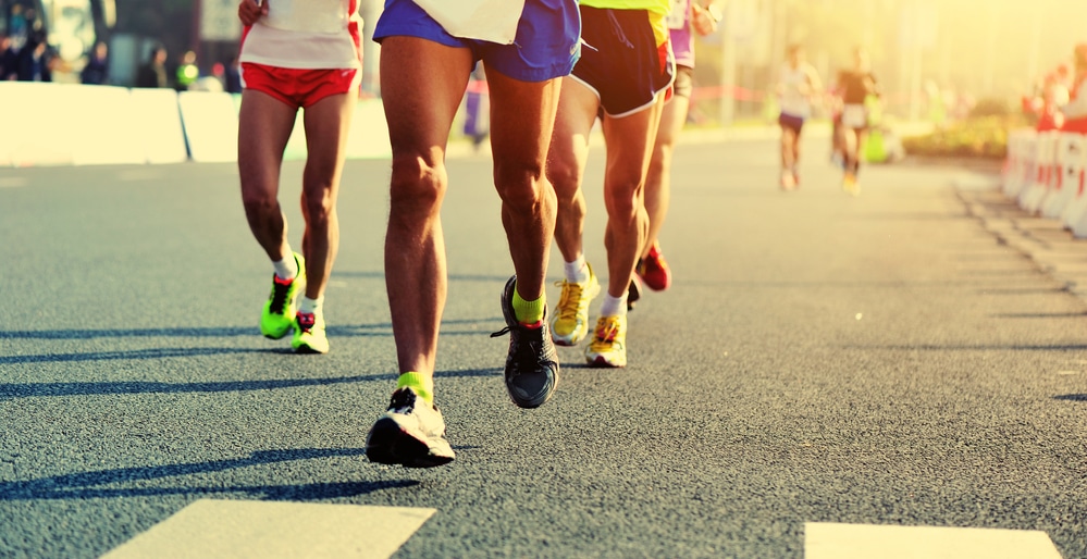 Pictured are the legs of a pack of four runners dressed in running shorts and running shoes, who are running on an asphalt road. The picture is possibly a 5K race in Frisco.