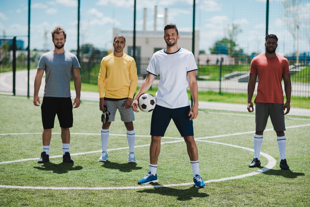 Four men wearing athletic gear are standing on a soccer field. The man in the center is holding a soccer ball on his right hip. The men may be part of one of the many Meetups taking place in Frisco.