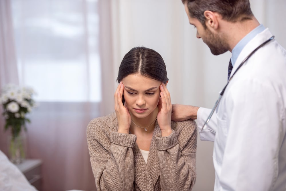 A dark-haired woman rubs her temples with her fingertips as she is consoled by a male doctor with short hair and a beard. She may suffer from high DHEA levels, a common problem among women.