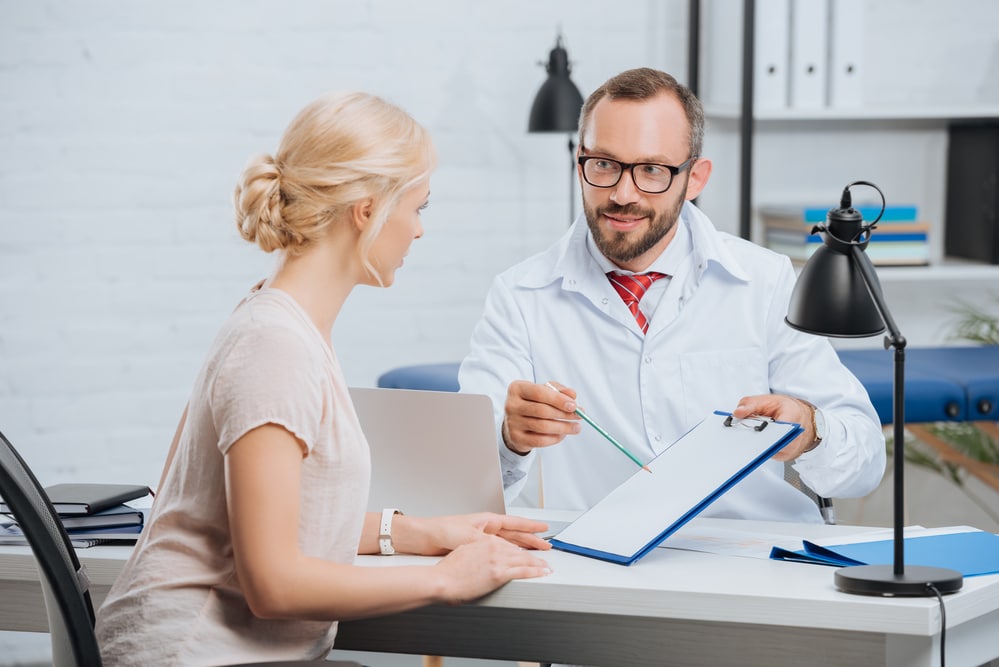 A blonde woman sits at an exam table, having a conversation with a bearded doctor who is showing her a chart on a clipboard. They are possibly discussing testosterone replacement therapy for women. 