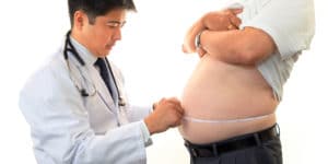A dark-haired doctor in a white lab coat measures the waist of a middle-aged man who is out of shape, possibly in preparation for a discussion on the affects of abdominal obesity on low testosterone levels.