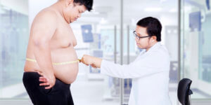 A male doctor uses a measuring tape to measure the stomach of an overweight male patient. The potential role of TRT in the prevention of diabetes may be discussed as part of his treatment plan.