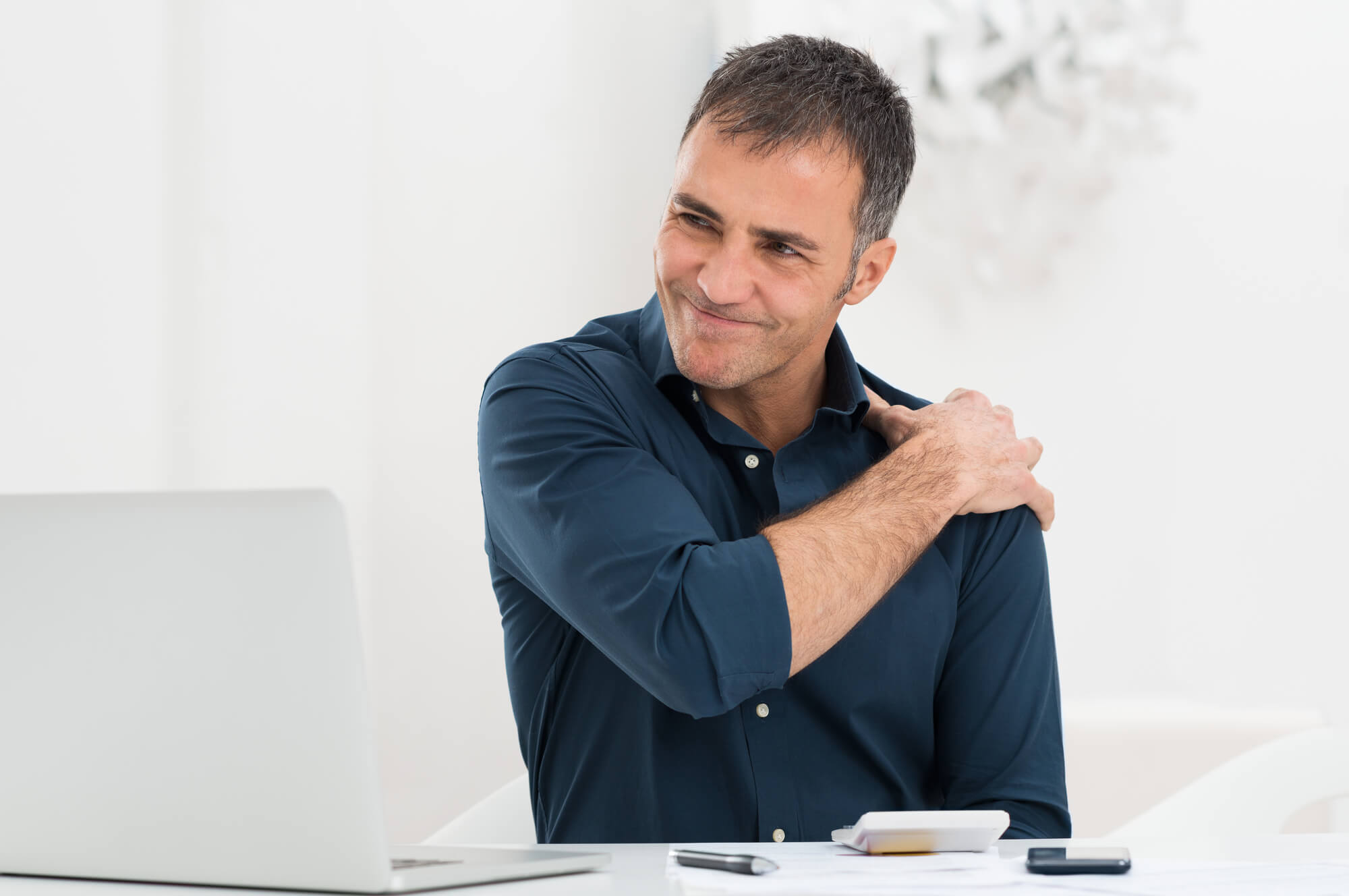 A middle-aged man in a dark blue shirt sits in front of his computer, massaging his shoulder and grimacing in pain. It's important that people in pain understand the connection between opioids and chronic low testosterone.