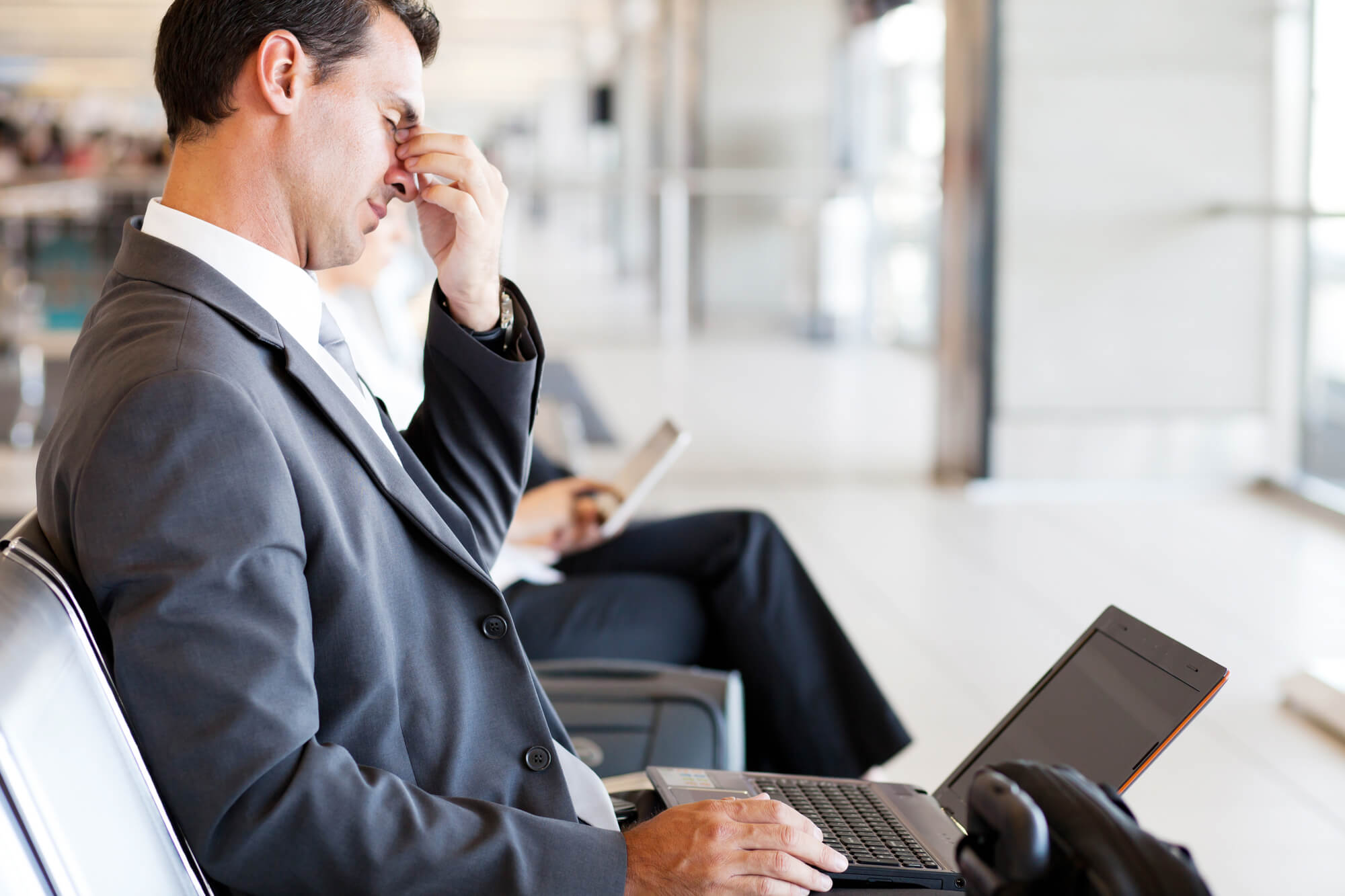 A tired businessman in a gray suit sits at an airport with a computer sitting on his lap. He is rubbing the bridge of his nose and looks fatigued. He may be suffering from the symptoms of male hypothyroidism.