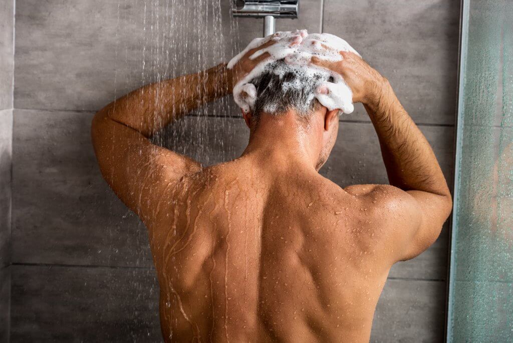 A rear view of a man's upper body as he shampoos his hair in the shower. He is likely unaware of the parabens in the shampoo that could contribute to low testosterone.