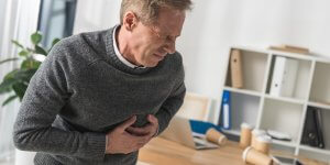 A man in a gray sweater clutches his chest, apparently from a heart attack. Research shows TRT is likely not the cause.