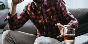 Man with a beard and plaid shirt reaches from the sofa to pick up a tumbler of alcohol, which can cause low testosterone when consumed to excess.