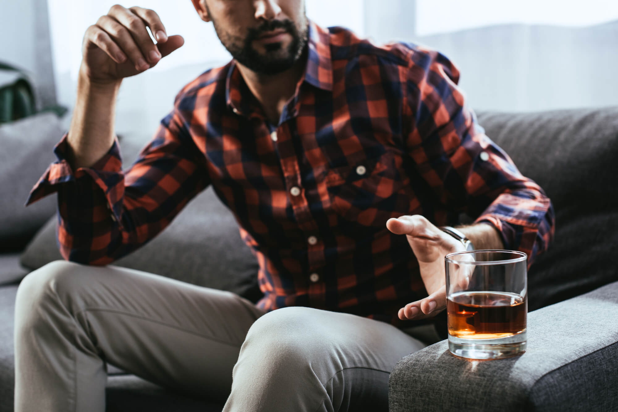 Man with a beard and plaid shirt reaches from the sofa to pick up a tumbler of alcohol, which can cause low testosterone when consumed to excess.