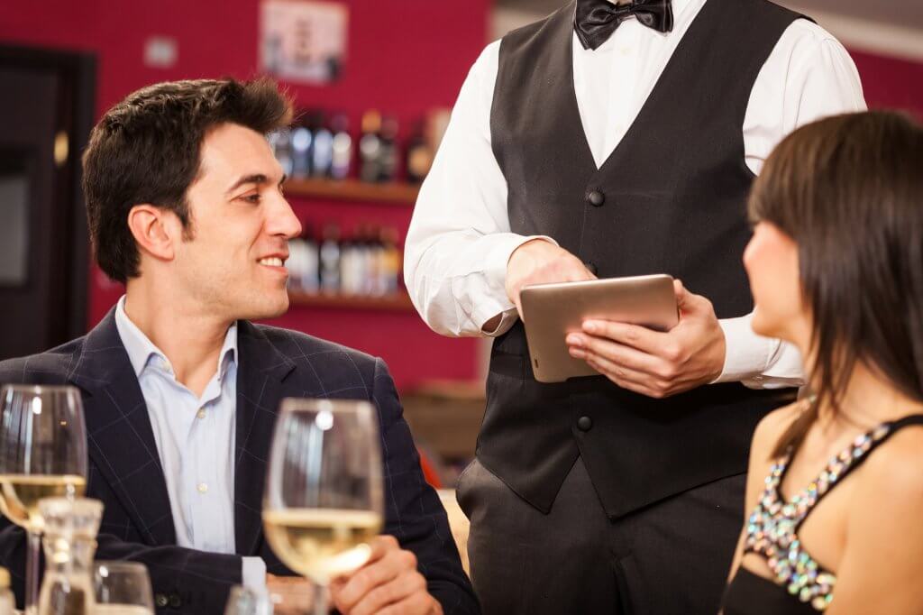A couple eat dinner at a fancy restaurant, served by a waiter wearing a white shirt and a black vest. The man may be concerned about the effect of reducing fat on his testosterone levels.