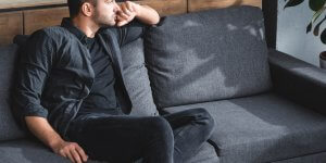 A man in dark clothes sits on a gray sofa. He's gazing out the window, possibly worried about low testosterone and its effect on his sex drive.