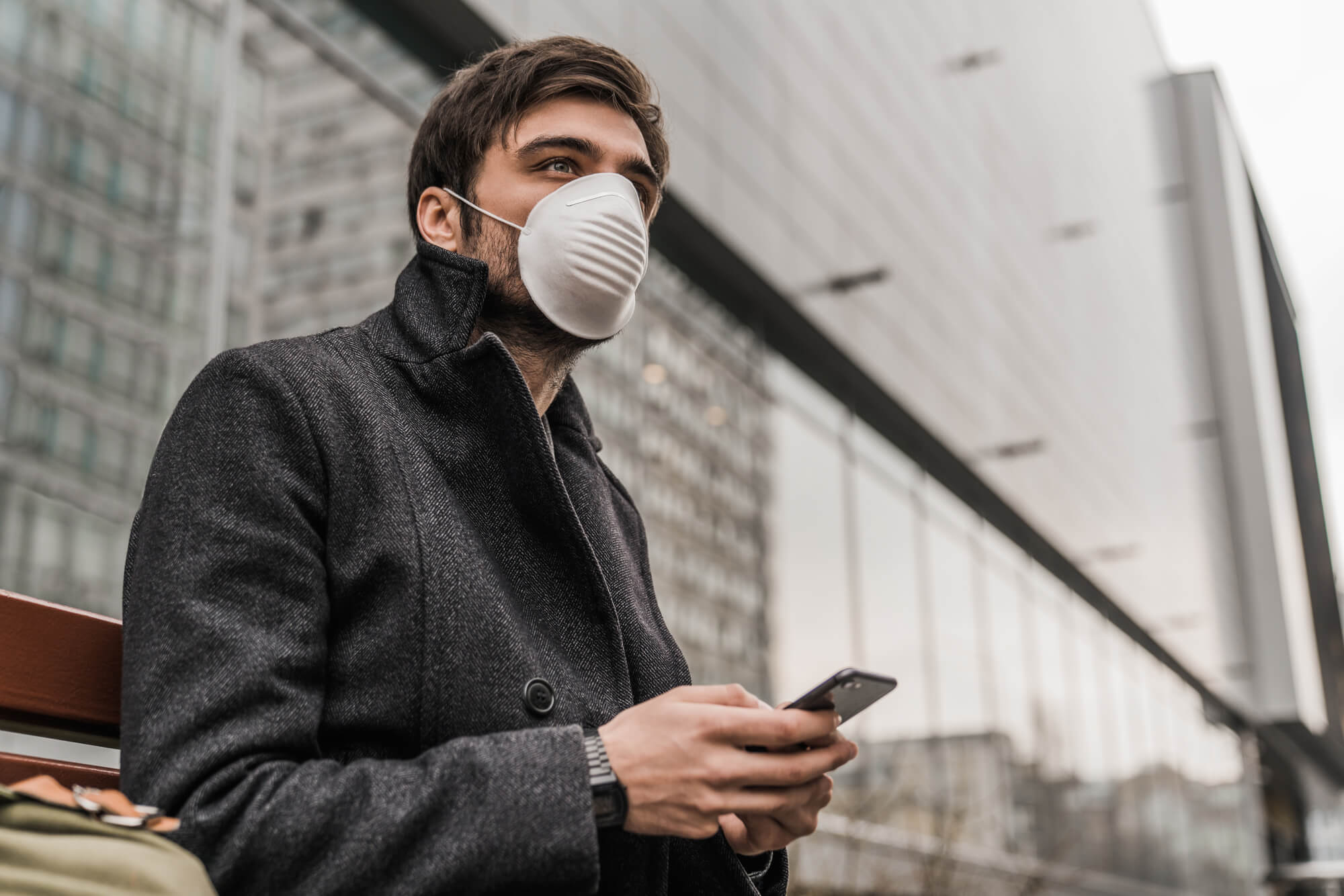A man in a gray jacket wears a protective face mask as he uses his cellular phone. He may be researching the connection between low testosterone and Covid 19.