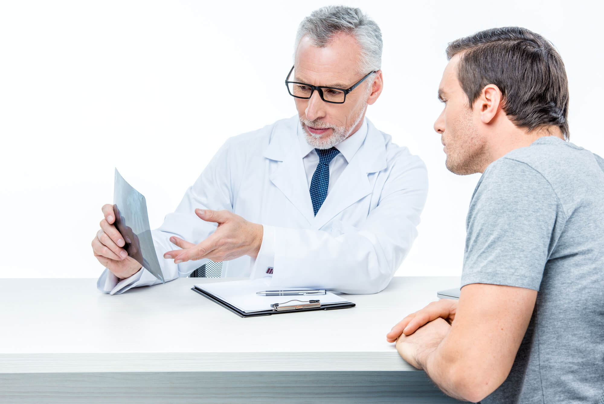 A male doctor wearing glasses is sitting at a white table with his male patient. The doctor is showing him a scan and explaining something, possibly how testosterone can affect prostate cancer.