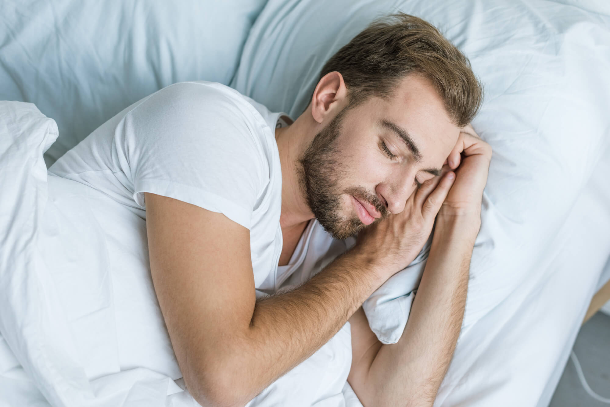 A man with a beard who wears a white t-shirt sleeps peacefully on white sheets.  Sleep and testosterone production have a strong connection.