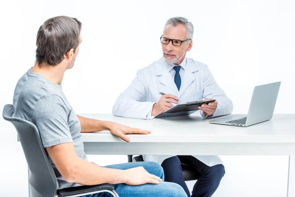 Doctor in a lab coat and tie sits at a white table with a male patient wearing a gray t-shirt, possibly discussing DHEA for men.