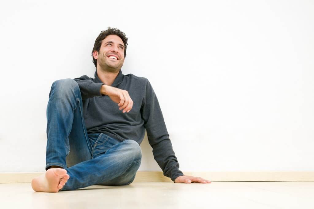 A barefoot man wearing worn jeans and a sweater leans against the wall. He may be experiencing the many TRT health benefits.