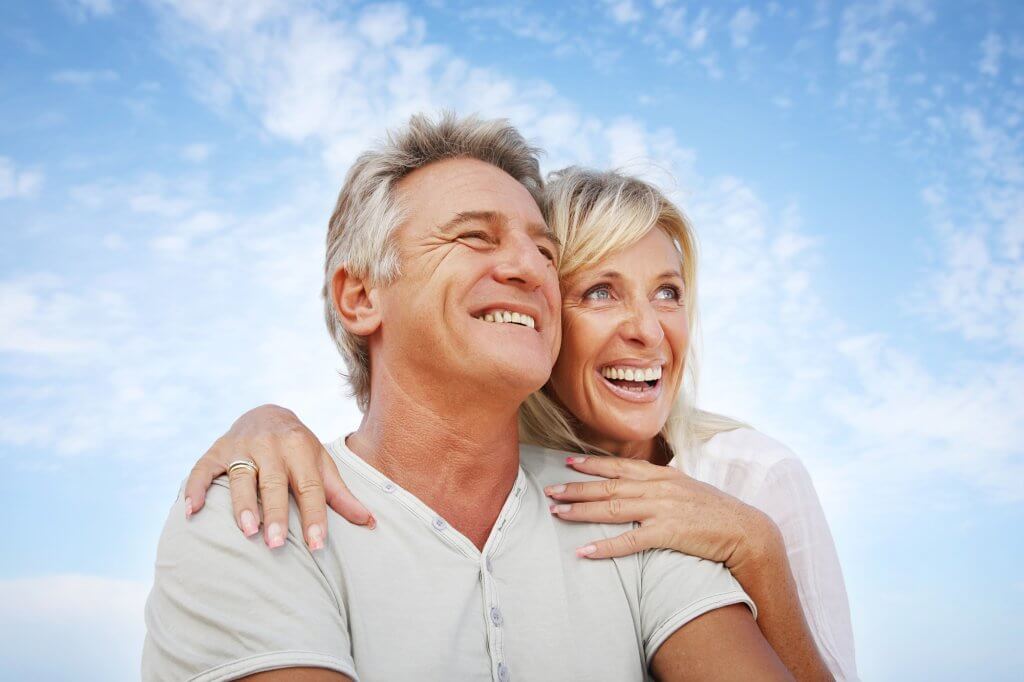 A middle-aged couple smile and embrace against a blue sky. Their lives have improved due to the TRT benefits to sexual performance.