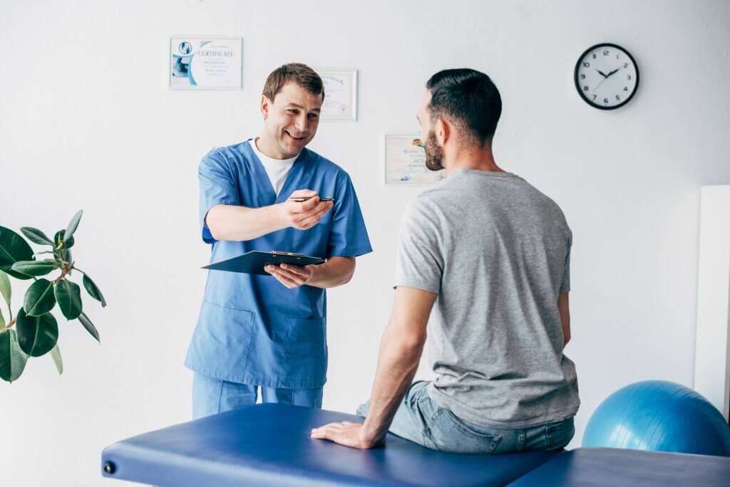 A doctor in scrubs talks to a male patient who sits on the exam table. Men's estrogen levels are something that should be discussed with your medical provider.