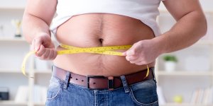 A man has pulled his white t-shirt up to reveal his belly, which slightly protrudes over his jeans and brown belt. He is measuring his waist and perhaps is concerned about his obesity risk.