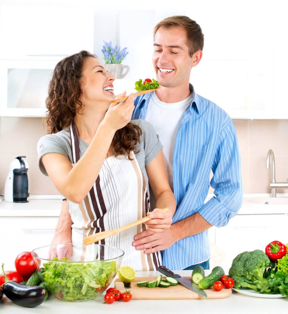 Una pareja feliz está cortando verduras para una ensalada. El hombre se coloca detrás de la mujer, y ella le da de comer ensalada por encima de su hombro izquierdo. Está comiendo sano esperando que una mejor dieta ayude a su testosterona.