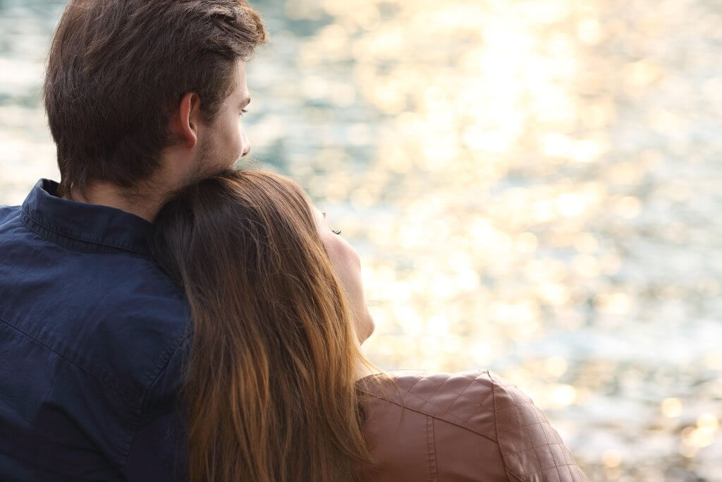 A woman sits with her head on a man's shoulder as they look at the water. There is a decline in male fertility that may complicate their relationship in the future.