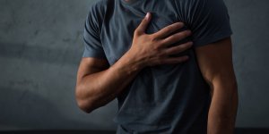 A man in a gray t-shirt clutches his left chest with his right hand. He may be concerned about a heart attack, but research shows this isn't a substantial trt risk.