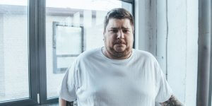 Overweight man in a white t-shirt looks concerned. Gynecomastia and obesity can occur with low testosterone.