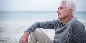 Silver haired man in a gray sweater sits on a beach. He may be suffering the affects of premature aging and low testosterone.