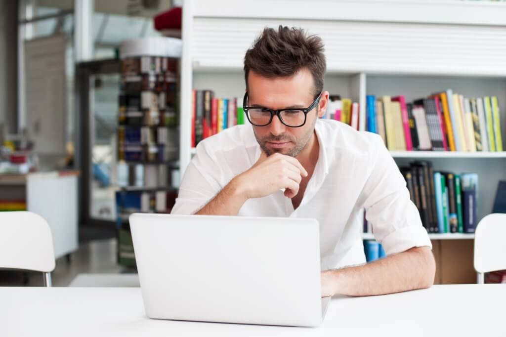A man wearing a white polo shirt and glasses does research on his laptop, perhaps wondering "What causes low testosterone?"