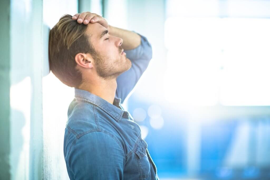 A man in a denim shirt leans back against a shiny wall. He is rubbing his forehead, perhaps worried about chronic Low T Health Risks.