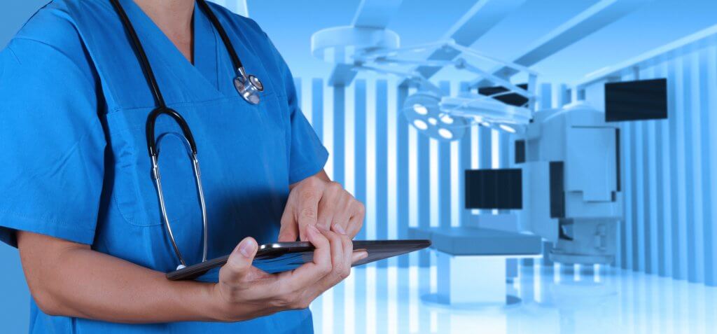 Doctor in blue scrubs stands n front of a medical device, possible used for administrating the digital rectal exam