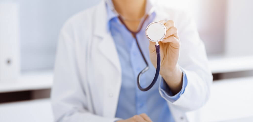 A female doctor in a blue blouse and white lab jacket is pictured from the shoulders to the waist, holding out a stethoscope with her left hand. Semaglutide may improve cardiac health.