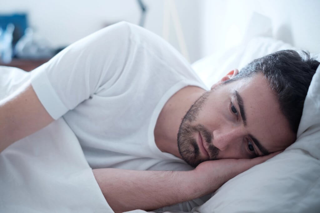 A man in a white t-shirt lies on his side in bed. He may need a sleep apnea mouth guard.