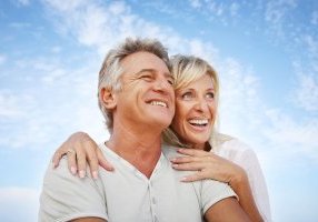 A middle-aged couple smile and embrace against a blue sky. Their lives have improved due to the TRT benefits to sexual performance.