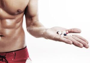 A muscular man's torso with his left arm outstretched can be seen. In his outstretched hand, he is holding a number of pills that could be testosterone boosters.