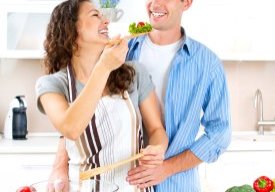 A happy couple are chopping vegetables for a salad. The man stands behind the woman, and she feeds him salad over her left shoulder. He's eating healthy hoping a better diet will help his testosterone.