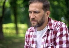 A man with a beard who is wearing a red plaid shirt over a white tee shirt looks concerned, perhaps learning that men need estrogen.